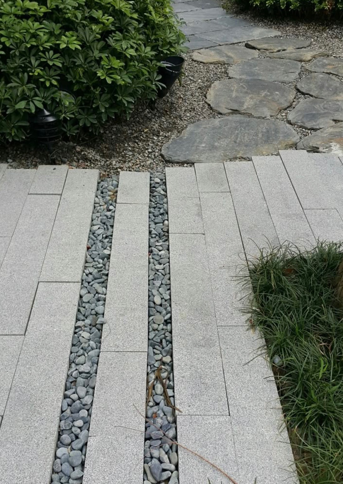 Szara ścieżka z prostokątnych płyt granitowych oddzielonych pasami drobnych kamieni, z roślinnością po bokach i kamienną ścieżką w tle.