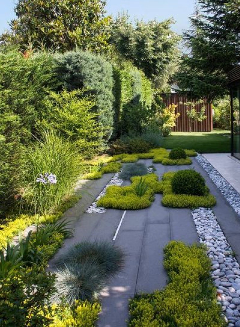 Nowoczesny ogród z szarymi płytami chodnikowymi, obłożony drobnymi kamieniami i ozdobnymi krzewami oraz trawami, z widoczną architekturą budynku w tle.