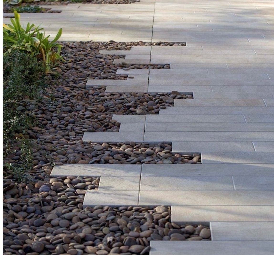 Ułożona ścieżka z jasnych, prostokątnych płyt chodnikowych z nieregularnymi przerwami wypełnionymi ciemnymi, okrągłymi kamieniami, obok rosną zielone rośliny.