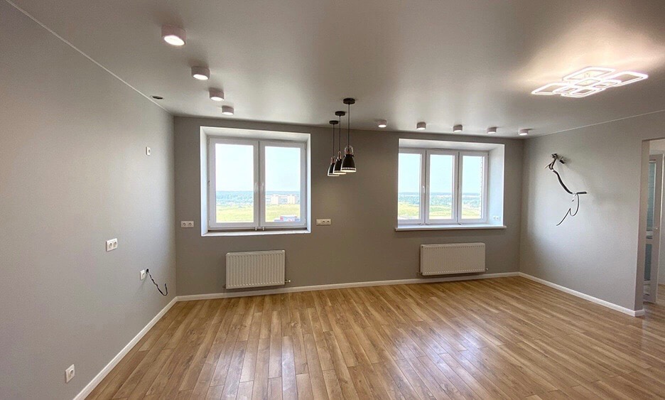 Jasne, przestronne wnętrze z parkietem w odcieniach brązu, szare ściany, dwa okna z widokiem na miasto, grzejniki pod oknami, nowoczesne oświetlenie sufitowe punktowe i wiszące, oraz instalacja...