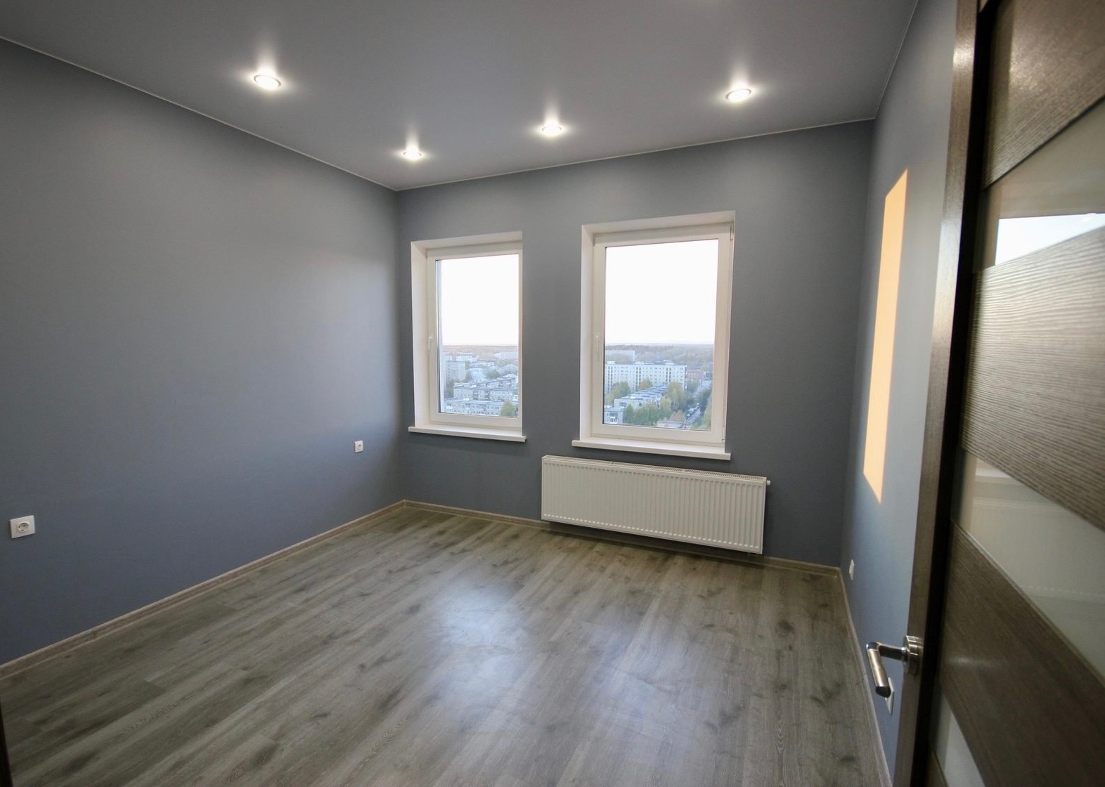 Pusty pokój w odcieniach szarości z panelami podłogowymi, białym grzejnikiem pod dwoma oknami z widokiem na miasto i nowoczesnymi drzwiami z elementami szkła.