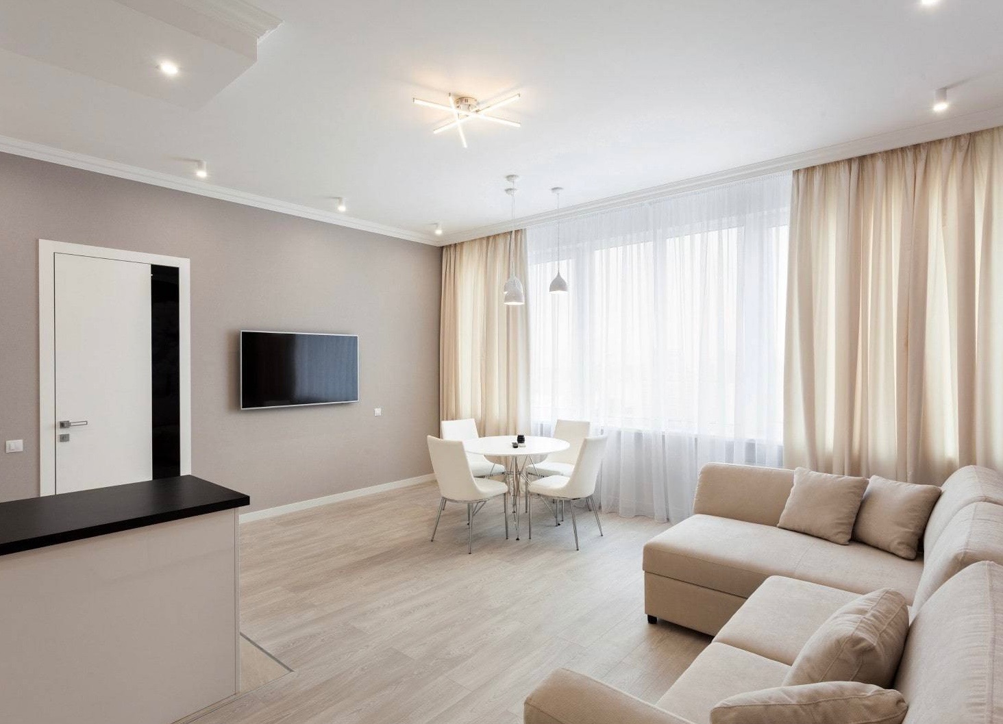 Jasne wnętrze salonu z beżową sofą narożną, okrągłym białym stołem z czterema krzesłami, telewizorem na ścianie i zasłonami w odcieniach beżu.