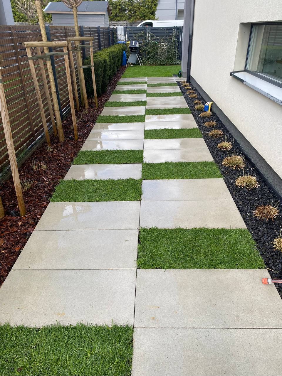 Ścieżka z jasnych płyt betonowych z zielonymi pasami trawy. Po bokach rabaty z korą i roślinami. Nowoczesny ogród z grillem w tle.