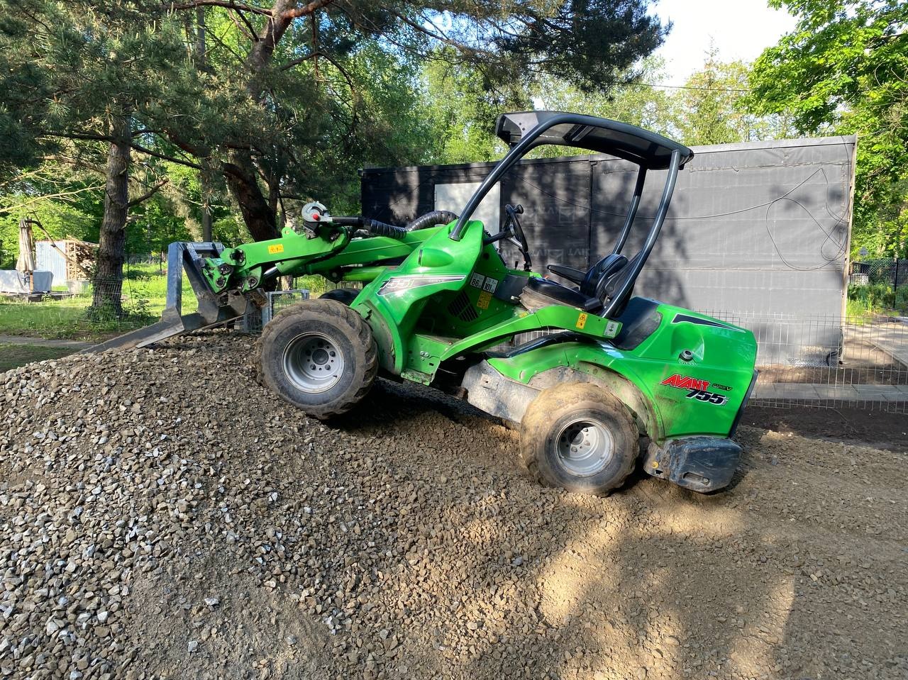 Zielona miniładowarka Avant 755 na żwirowym podłożu, gotowa do pracy przy niwelacji terenu. W tle widoczne drzewa i fragment ogrodzenia.