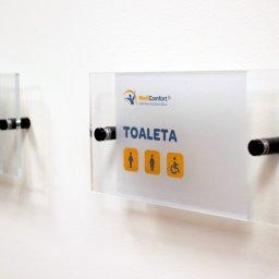 AGENCJA REKLAMY FUX SP.Z O.O. - Szklane tabliczki informacyjne z logo i piktogramami, przymocowane do ściany za pomocą metalowych dystansów, wskazujące na toaletę i gabinet.