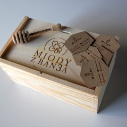 AGENCJA REKLAMY FUX SP.Z O.O. - Drewniana skrzynka z wygrawerowanym logo 'Miody z Ran3a' oraz drewnianym nabierakiem do miodu i stosem wizytówek z motywem pszczoły.
