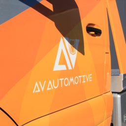 AGENCJA REKLAMY FUX SP.Z O.O. - Pomarańczowy samochód dostawczy z białym logo 'AV AUTOMOTIVE' i elementami graficznymi, zaparkowany na tle szarej ściany.