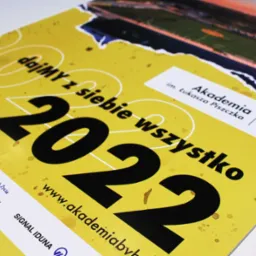 Żółty kalendarz na rok 2022 z hasłem 'dajMY z siebie wszystko', logotypami sponsorów i zdjęciem stadionu, promujący Akademię im. Łukasza Piszczka z logiem BVB 09.