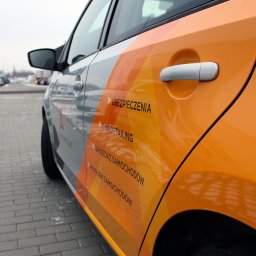 AGENCJA REKLAMY FUX SP.Z O.O. - Fragment samochodu z oklejeniem reklamowym w odcieniach szarości i pomarańczy, prezentującym usługi: ubezpieczenia, car detailing, sprzedaż i wynajem samochodów.
