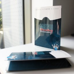 AGENCJA REKLAMY FUX SP.Z O.O. - Dwa menu bowlingowe z grafiką kuli i kręgli, jedno stojące, drugie leżące na białym blacie, z napisem 'MENU Bowling Casamento' i hasłem 'Wybierz Zamów Zjedz'.