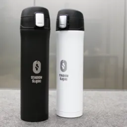 Dwie metalowe butelki termiczne, czarna i biała, z nadrukowanym logo i napisem 'Stadion Śląski', stojące na szarej powierzchni z metalowym tłem.