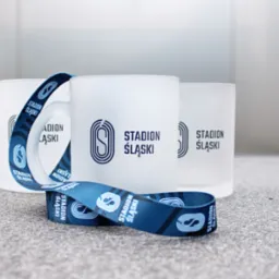 Trzy matowe szklanki i smycz z nadrukiem logo Stadionu Śląskiego, ułożone na szarym blacie, w tle jasnoszara ściana.