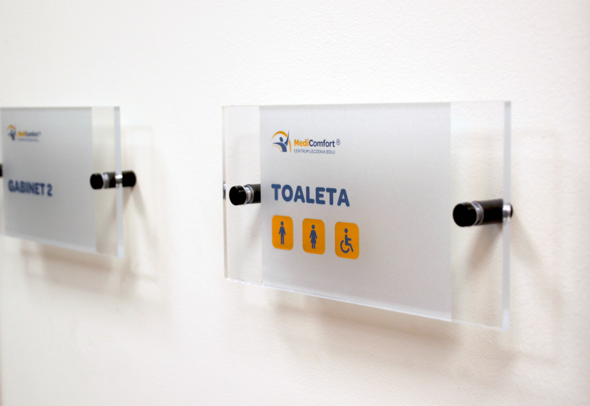 Szklane tabliczki informacyjne z logo i piktogramami, przymocowane do ściany za pomocą metalowych dystansów, wskazujące na toaletę i gabinet.