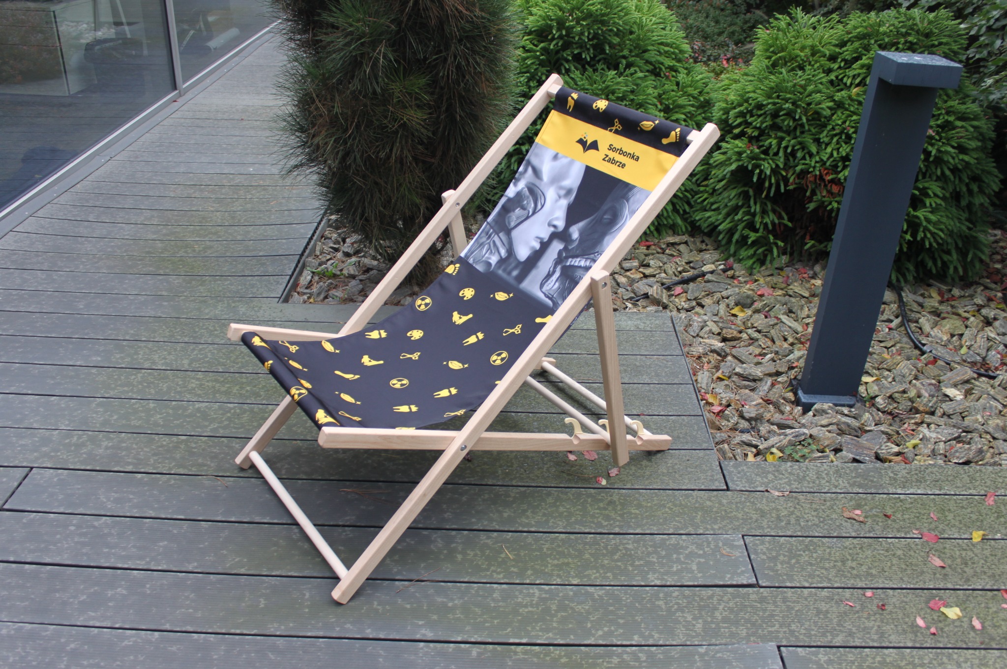 Drewniany leżak z nadrukiem logo i grafiką firmy na tle drewnianego tarasu i zieleni.