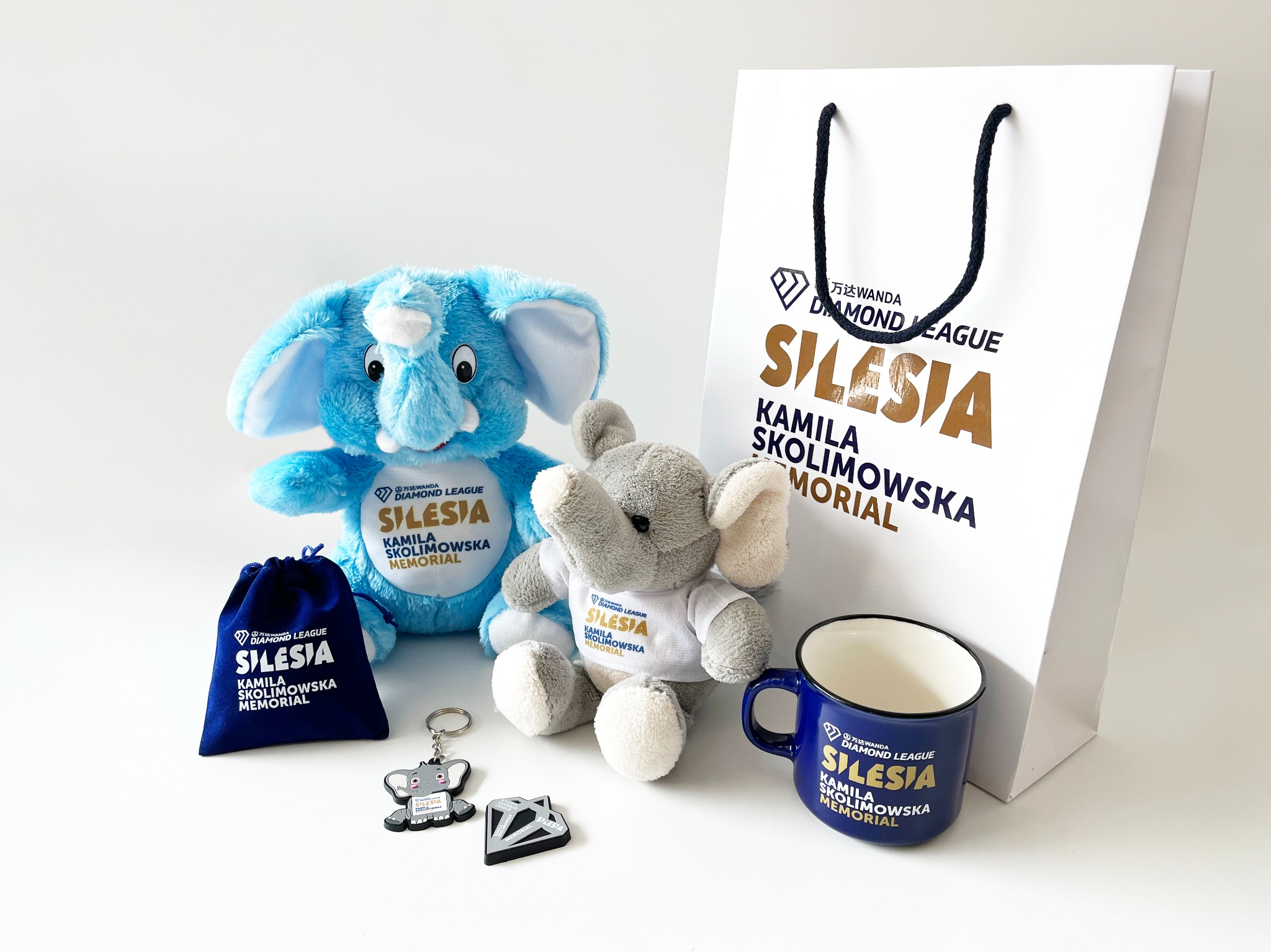 Zestaw gadżetów promocyjnych Wanda Diamond League Silesia Kamila Skolimowska Memorial: pluszowe słonie w koszulkach, brelok, woreczek, torba papierowa i emaliowany kubek na białym tle.