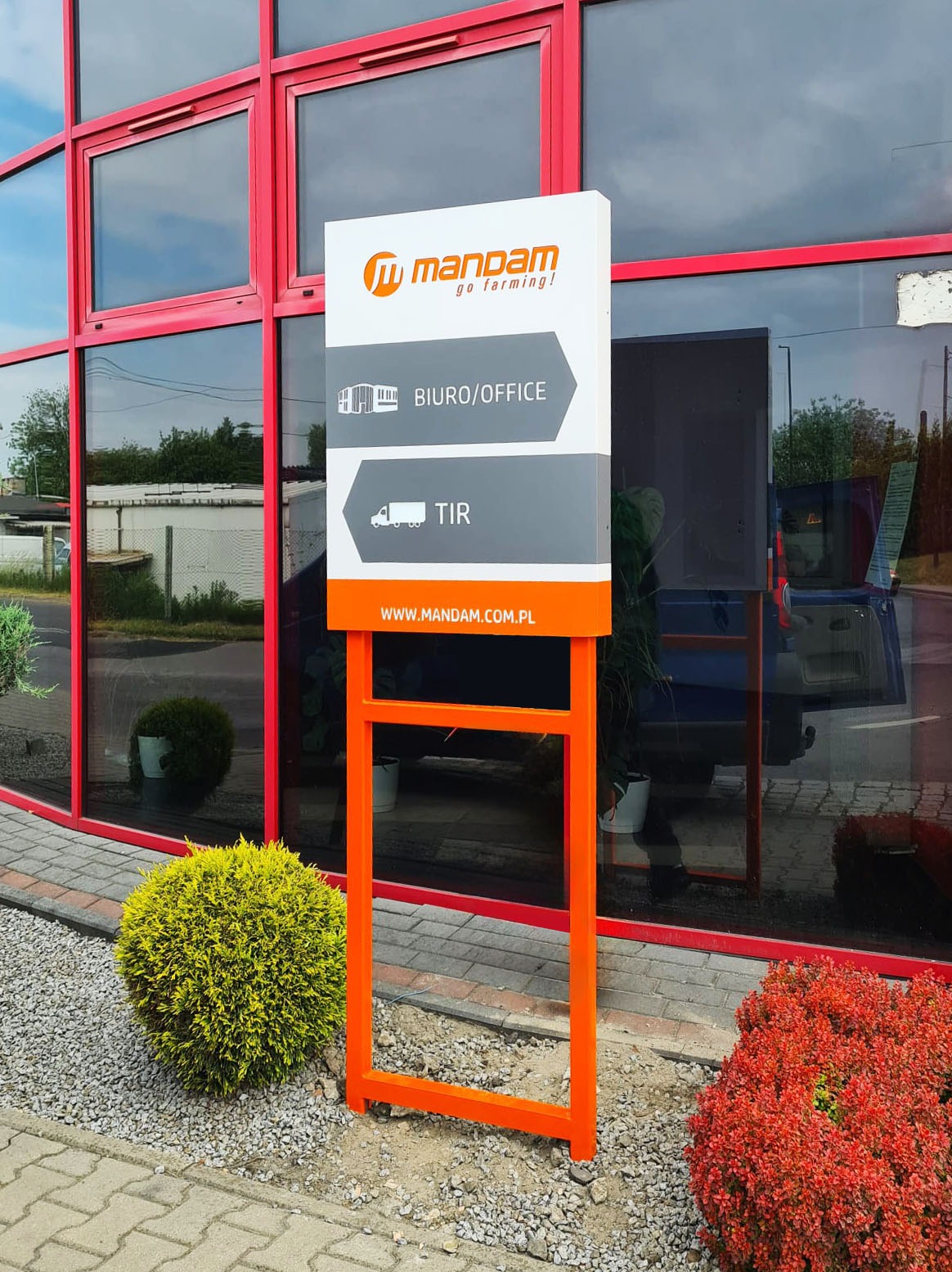 Zewnętrzny pylon informacyjny firmy Mandam z pomarańczową ramą, wskazujący kierunki do biura i działu TIR, umieszczony przed budynkiem z czerwonymi ramami okiennymi.