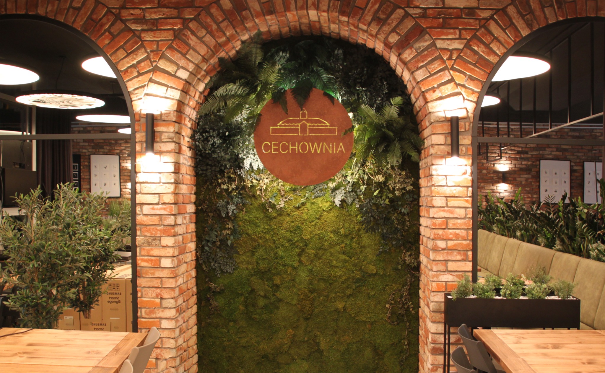 Wnętrze restauracji z ceglanym łukiem, zieloną ścianą z mchu i roślin, oraz okrągłym szyldem z napisem CECHOWNIA i graficznym symbolem budynku.
