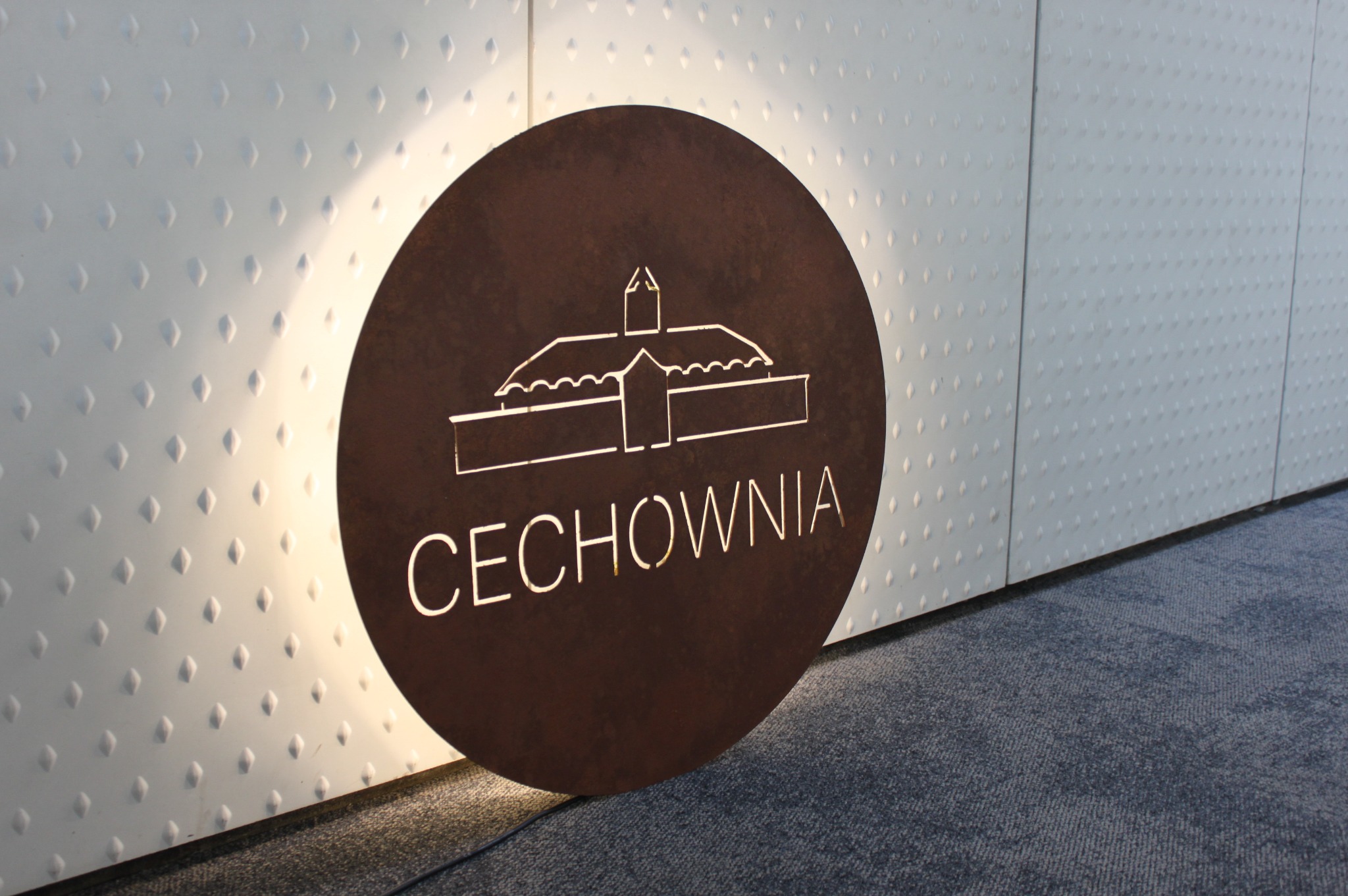 Rustykalny, okrągły szyld z wyciętym logo budynku i napisem 'CECHOWNIA', podświetlony od tyłu, ustawiony na szarym dywanie na tle białej, teksturowanej ściany.