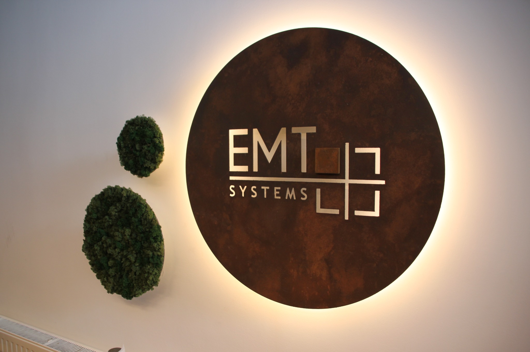 Podświetlane logo firmy EMT Systems na okrągłej, rdzawobrązowej płycie, ozdobione dwoma kulami z mchu na białej ścianie.
