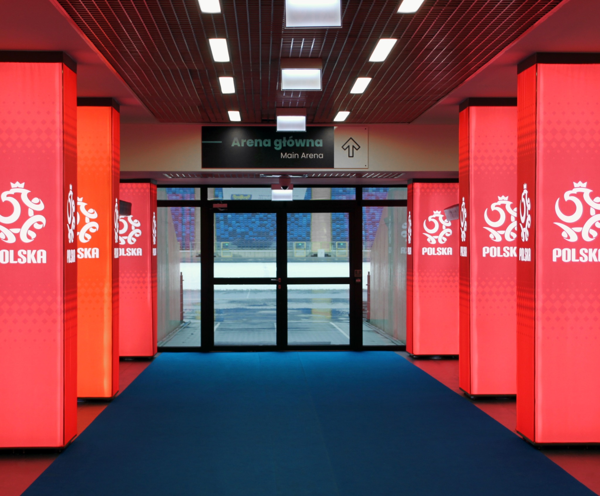 Wnętrze areny sportowej z rzędem podświetlanych, czerwonych kolumn z logo i napisem 'Polska', prowadzących do szklanych drzwi wejściowych z widokiem na trybuny.