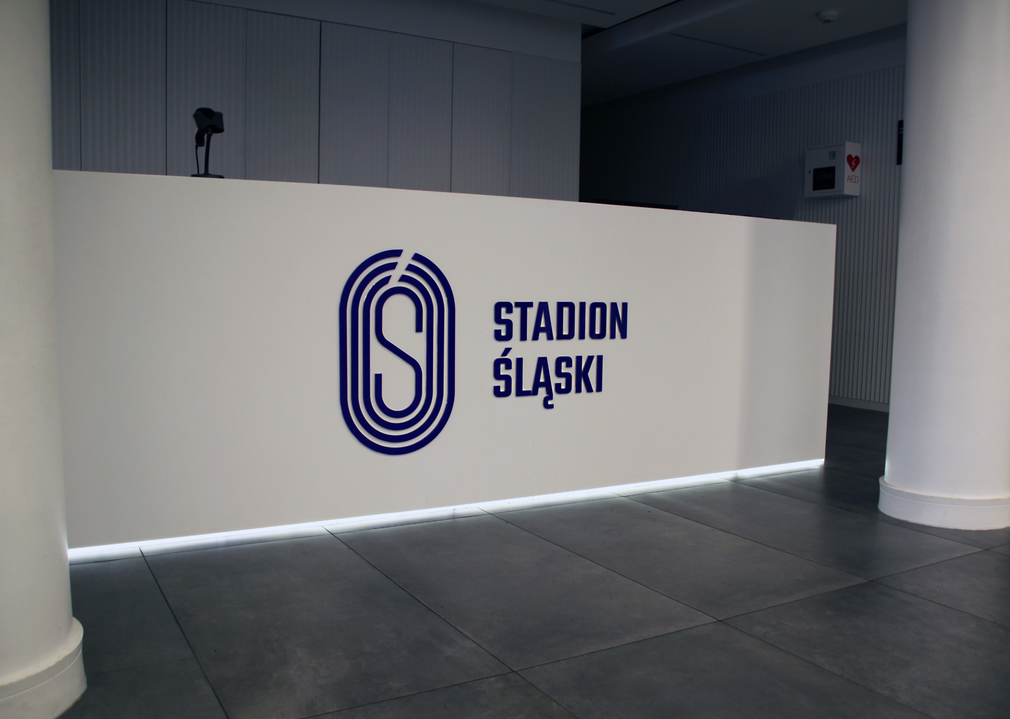 Recepcja z logo Stadionu Śląskiego w niebieskim kolorze, podświetlona listwą LED, widoczny automat AED na ścianie w tle.