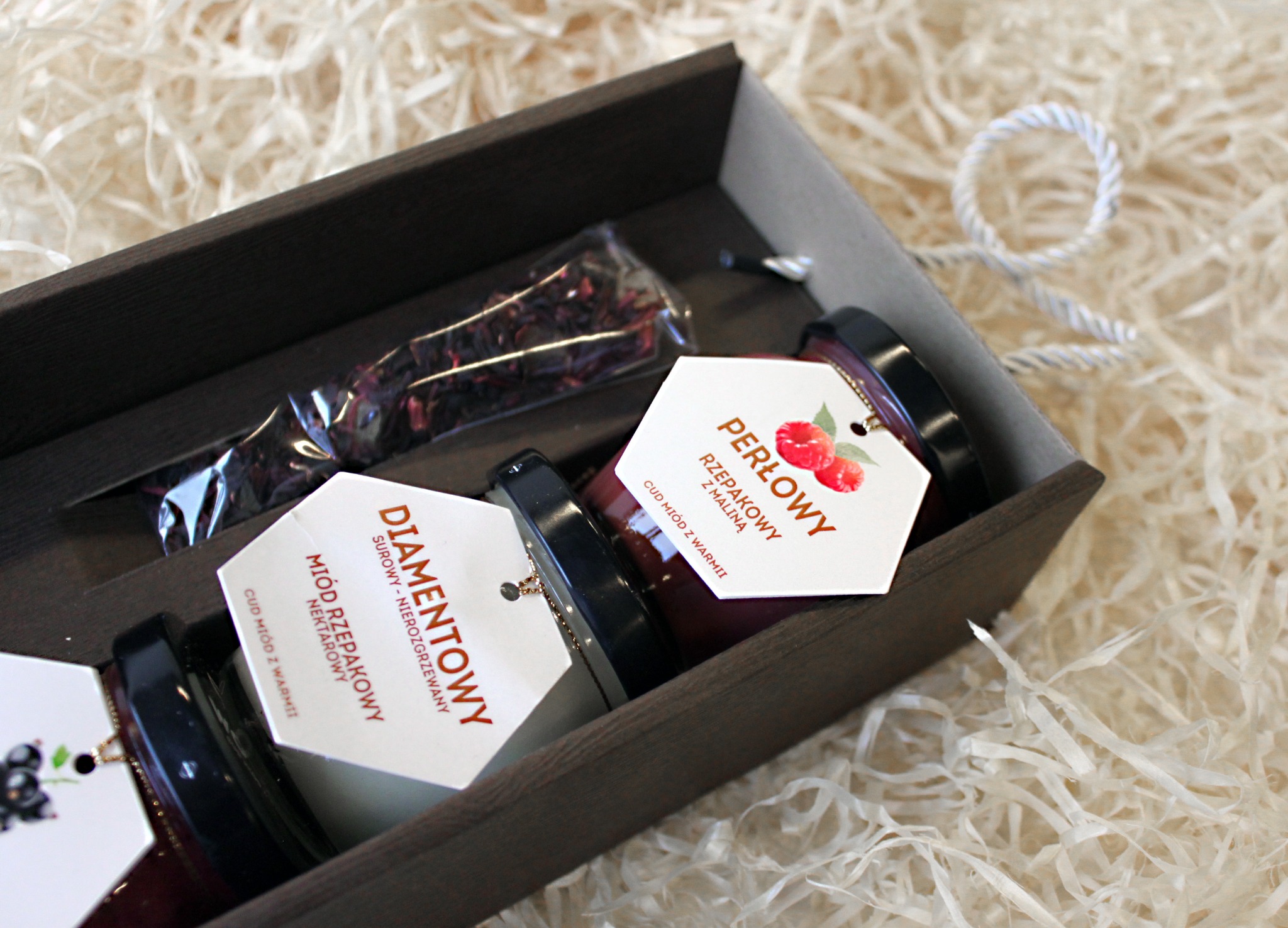 Zestaw prezentowy: dwa słoiki miodu rzepakowego z etykietami 'Perłowy' (z maliną) i 'Diamentowy' oraz paczka suszonych kwiatów w ciemnobrązowym drewnianym pudełku wypełnionym białym siankiem...