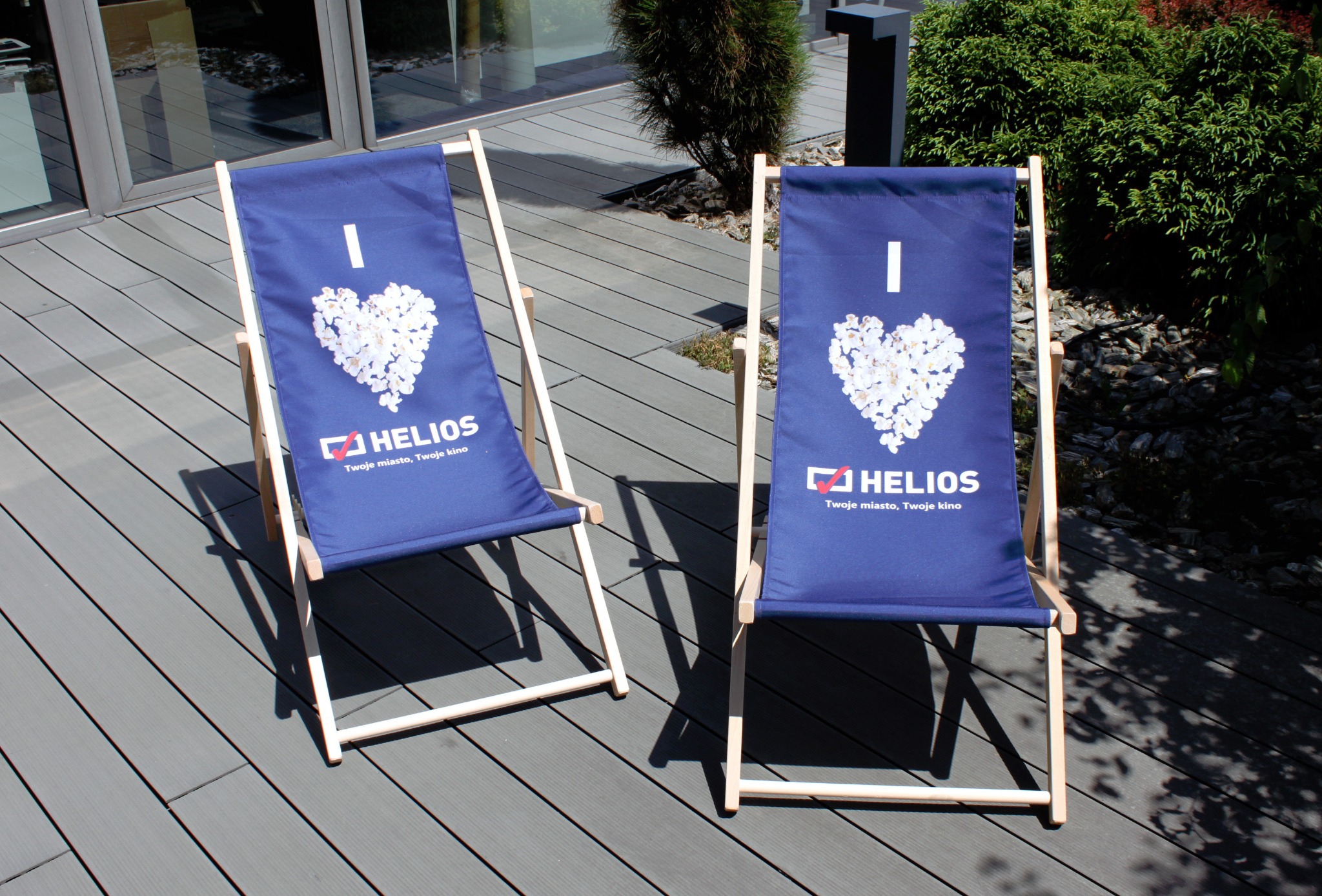 Dwa leżaki plażowe z drewnianą ramą i niebieskim płótnem z nadrukiem 'I serce z popcornu HELIOS Twoje miasto, Twoje kino' stoją na drewnianym tarasie na zewnątrz budynku.