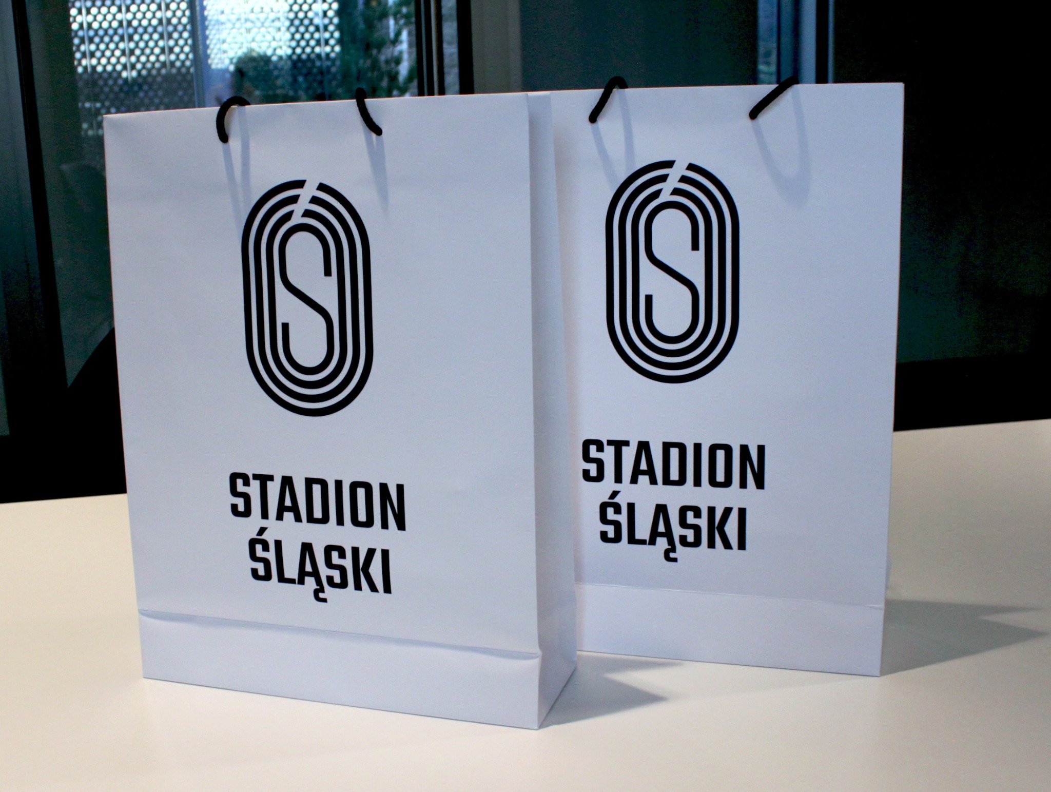 Dwie białe torby papierowe z czarnym nadrukiem logo i napisem 'Stadion Śląski', ustawione na jasnym blacie.