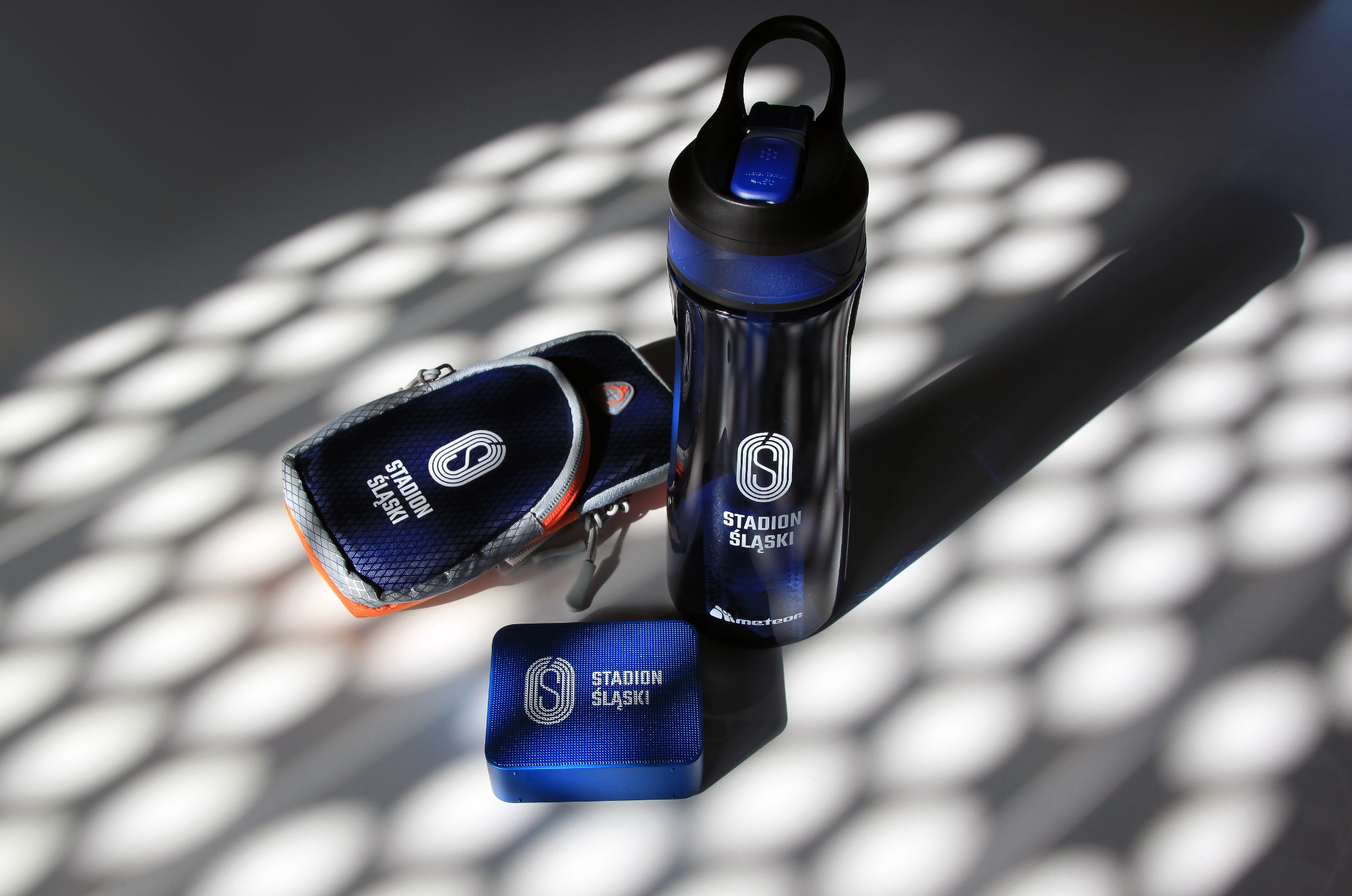 Zestaw promocyjny Stadionu Śląskiego: bidon, etui i powerbank z logo, na tle wzoru cienia.