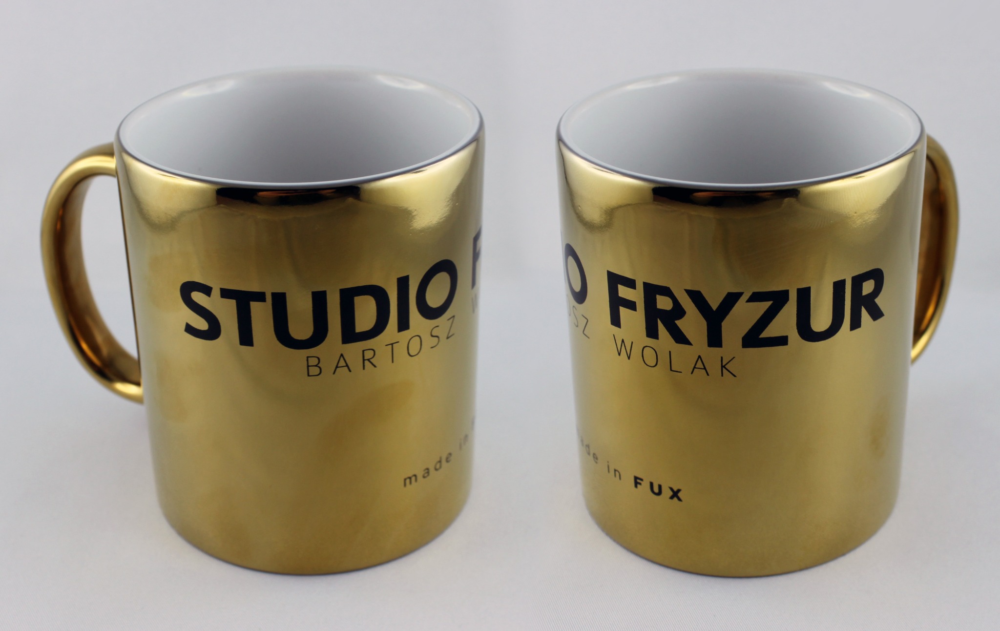 Dwa złote kubki z nadrukowanymi nazwami salonów fryzjerskich: 'Studio Bartosz' i 'O Fryzur Wolak', ustawione obok siebie na białym tle.