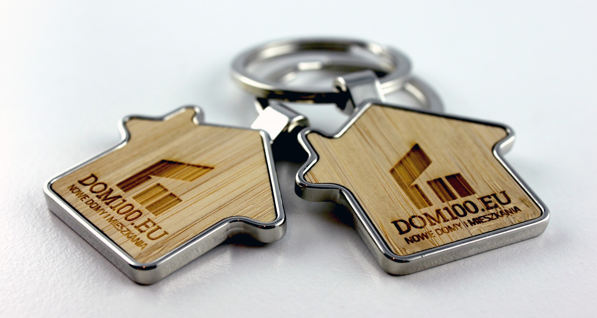 Dwa drewniane breloczki w kształcie domu z metalową obwódką i logo DOM100.EU, leżące na białym tle, z metalowym kółkiem do kluczy w tle.
