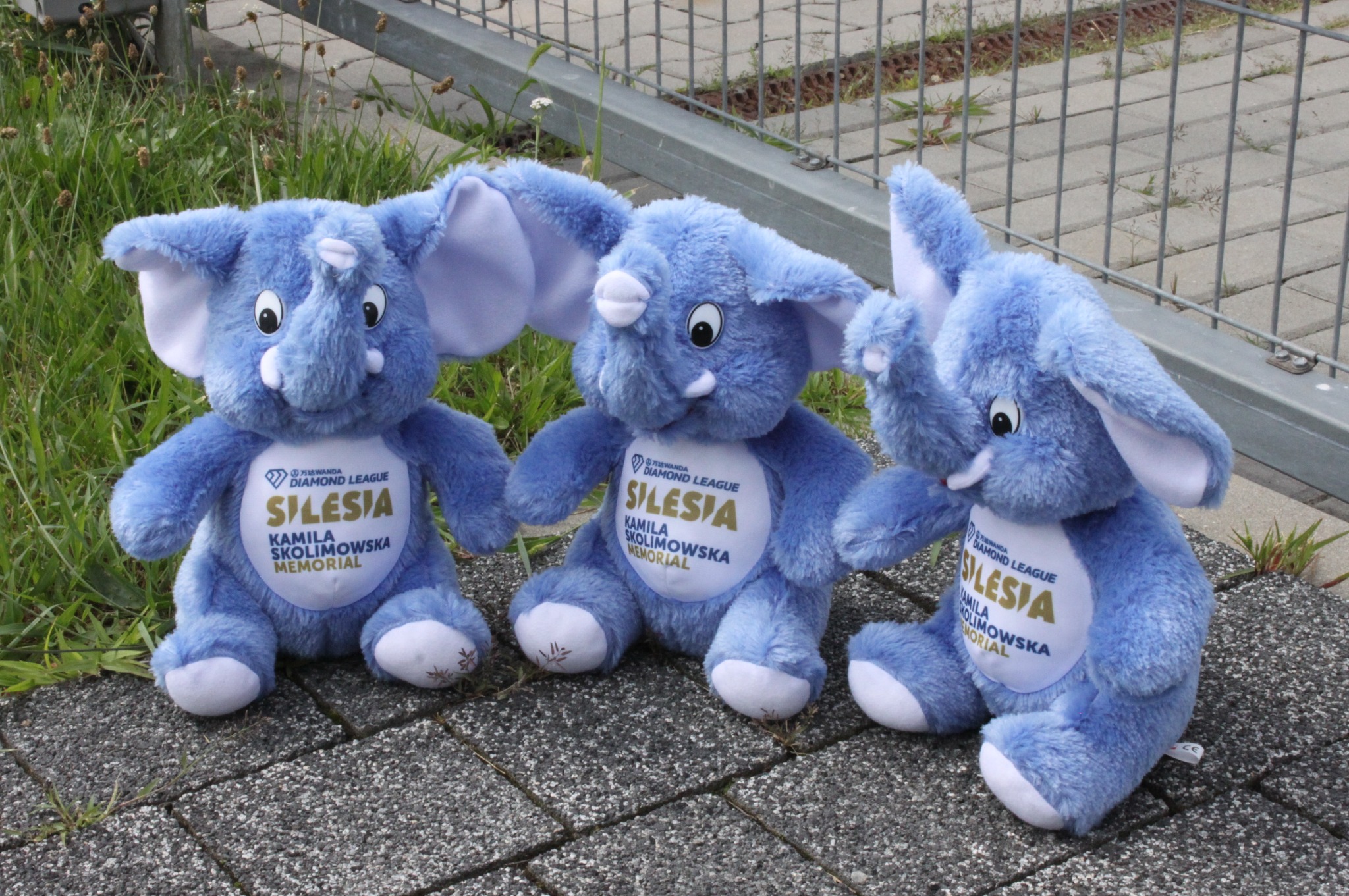 Trzy niebieskie pluszowe słonie z logo Wanda Diamond League Śląsk Kamila Skolimowska Memorial, ustawione na chodniku w Gliwicach, obok metalowego ogrodzenia i trawy.
