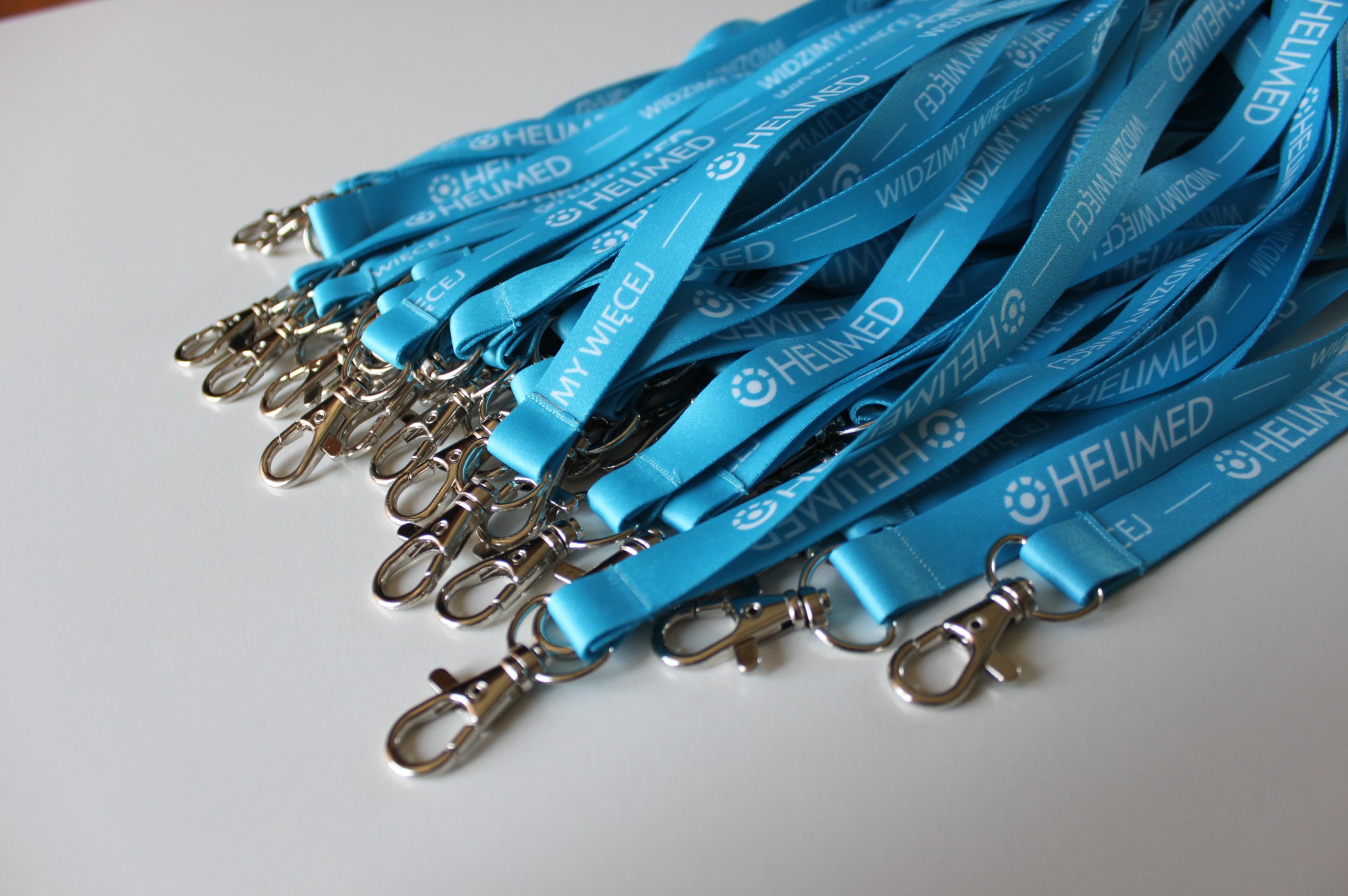 Stos niebieskich smyczy z karabińczykami, z nadrukowanym logo i napisem 'HELIMED - WIDZIMY WIĘCEJ', leżących na białym tle.