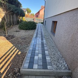 “ J&P.B ” Mycie dachów i elewacji - Ścieżka z kostki brukowej w odcieniach szarości prowadząca wzdłuż ściany domu z oknem, obok rabata wysypana kamieniami i krzewami, w tle inne domy.