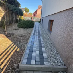 Ścieżka z kostki brukowej w odcieniach szarości prowadząca wzdłuż ściany domu z oknem, obok rabata wysypana kamieniami i krzewami, w tle inne domy.