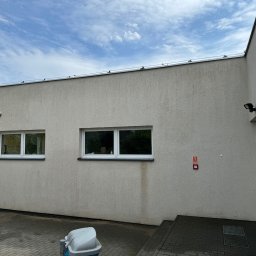 “ J&P.B ” Mycie dachów i elewacji - Jasna elewacja budynku z dwoma oknami i widocznymi śladami zacieków poniżej jednego z nich, alarm przeciwpożarowy na ścianie, kostka brukowa na dziedzińcu