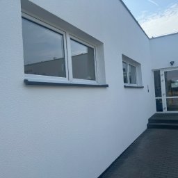 “ J&P.B ” Mycie dachów i elewacji - Świeżo pomalowana, biała elewacja budynku z dwoma oknami i wejściem, widoczna faktura tynku baranka.