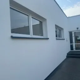 Świeżo pomalowana, biała elewacja budynku z dwoma oknami i wejściem, widoczna faktura tynku baranka.