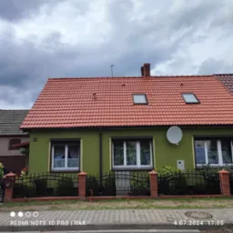 Dom jednorodzinny z czerwoną dachówką, zieloną elewacją, numerem 10 i anteną satelitarną na ścianie, sfotografowany w pochmurny dzień.