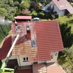 “ J&P.B ” Mycie dachów i elewacji - Pracownik na podnośniku koszowym czyści dach z czerwonej dachówki wodą pod ciśnieniem, widoczny efekt czyszczenia - część dachu umyta, część zabrudzona, dom z jasną elewacją w tle.