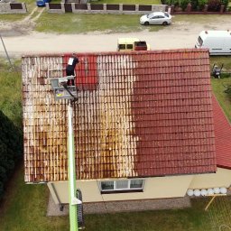 “ J&P.B ” Mycie dachów i elewacji - Pracownik na podnośniku koszowym czyści połowę dachu z czerwonej dachówki, widoczny efekt przed i po czyszczeniu, otoczenie to trawnik, dom i droga.