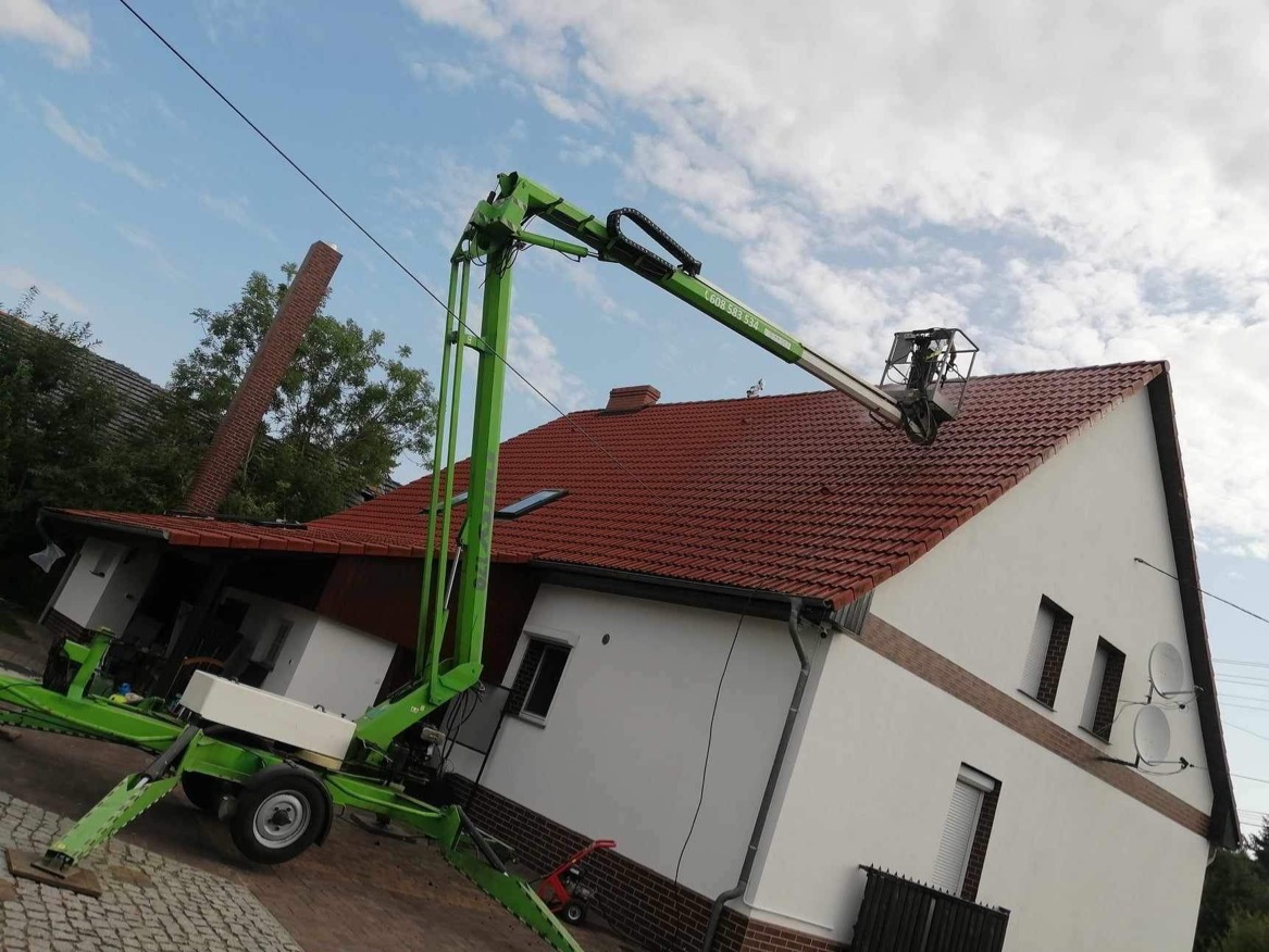 Prace na dachu domu jednorodzinnego w Rzepinie z wykorzystaniem podnośnika koszowego. Widoczny fragment komina i biała elewacja budynku.