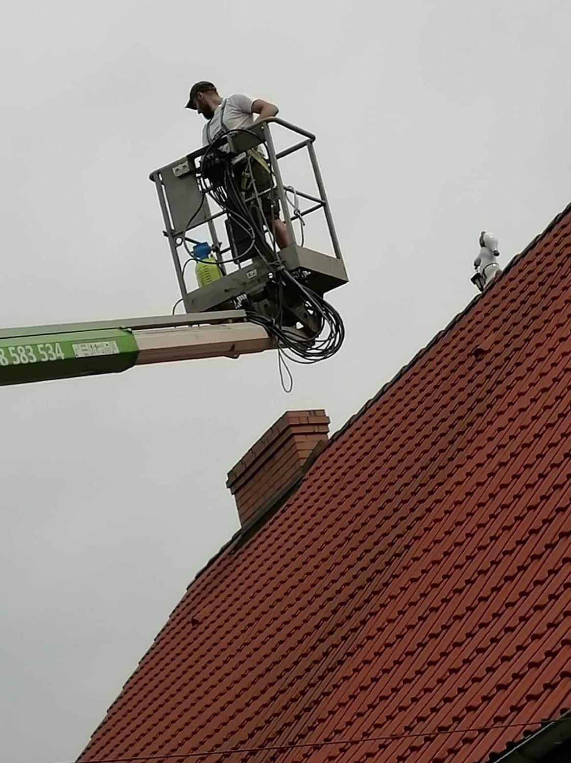 Pracownik na podnośniku koszowym przy dachu z czerwonej dachówki, obok komin i ozdobny element na dachu w kształcie psa.