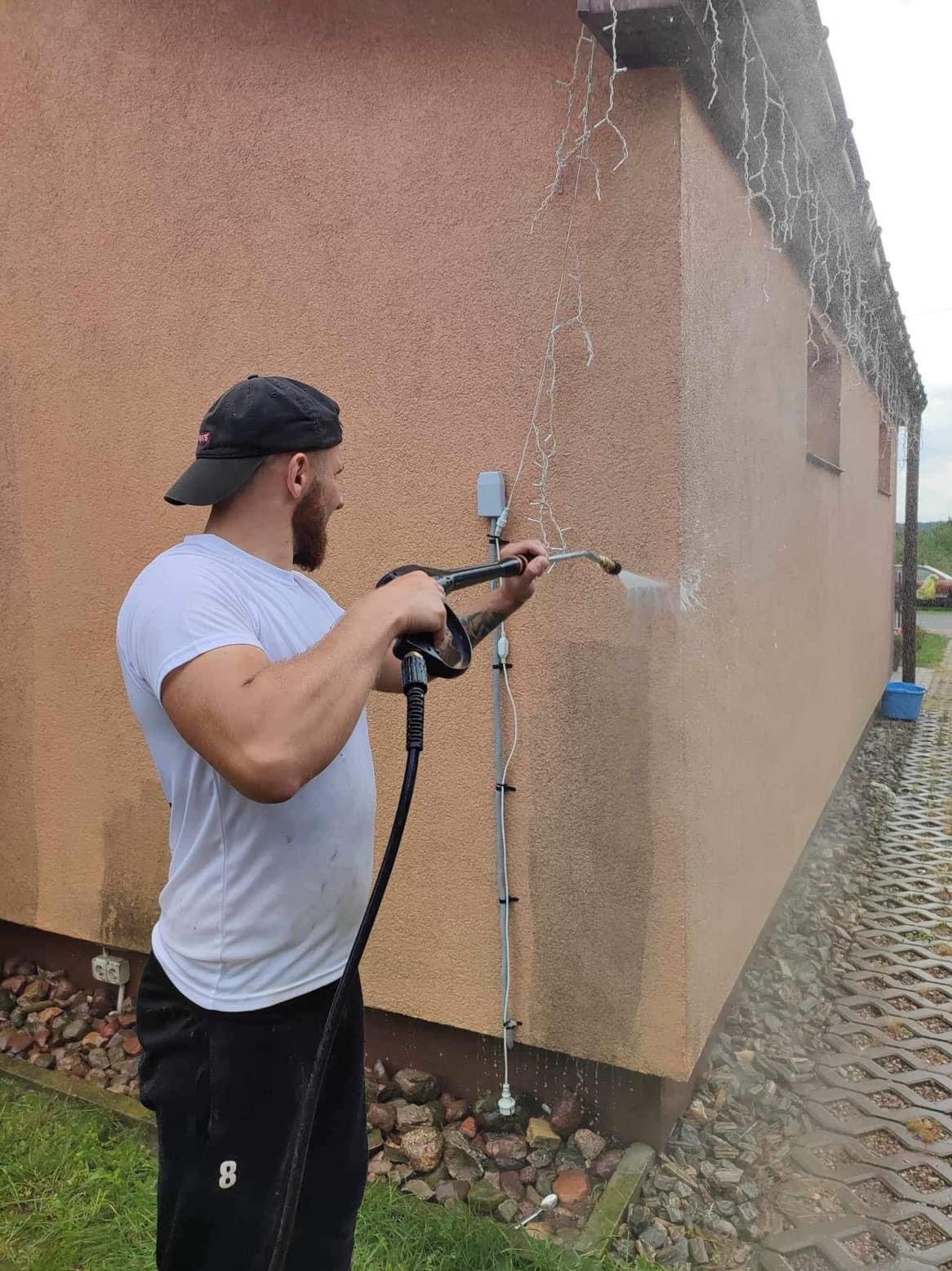 Mężczyzna w czapce i białej koszulce czyści elewację domu myjką ciśnieniową, w tle widoczne lampki świąteczne pod dachem i kostka brukowa.