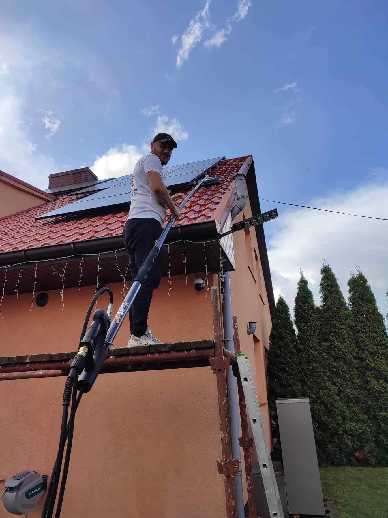 Mężczyzna w czapce i białej koszulce, stojący na rusztowaniu, czyści dach z paneli słonecznych za pomocą długiej szczotki teleskopowej, widoczny fragment rynny i elewacji budynku z oknami, w tle...