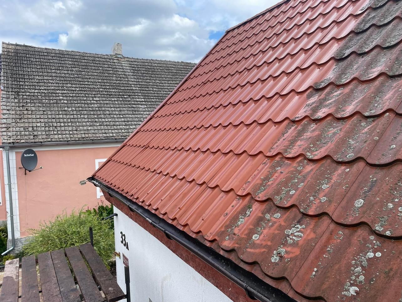 Częściowo wyczyszczony czerwony dach z blachodachówki, widoczne zanieczyszczenia biologiczne i osady przed i po czyszczeniu, w tle budynek z anteną satelitarną.