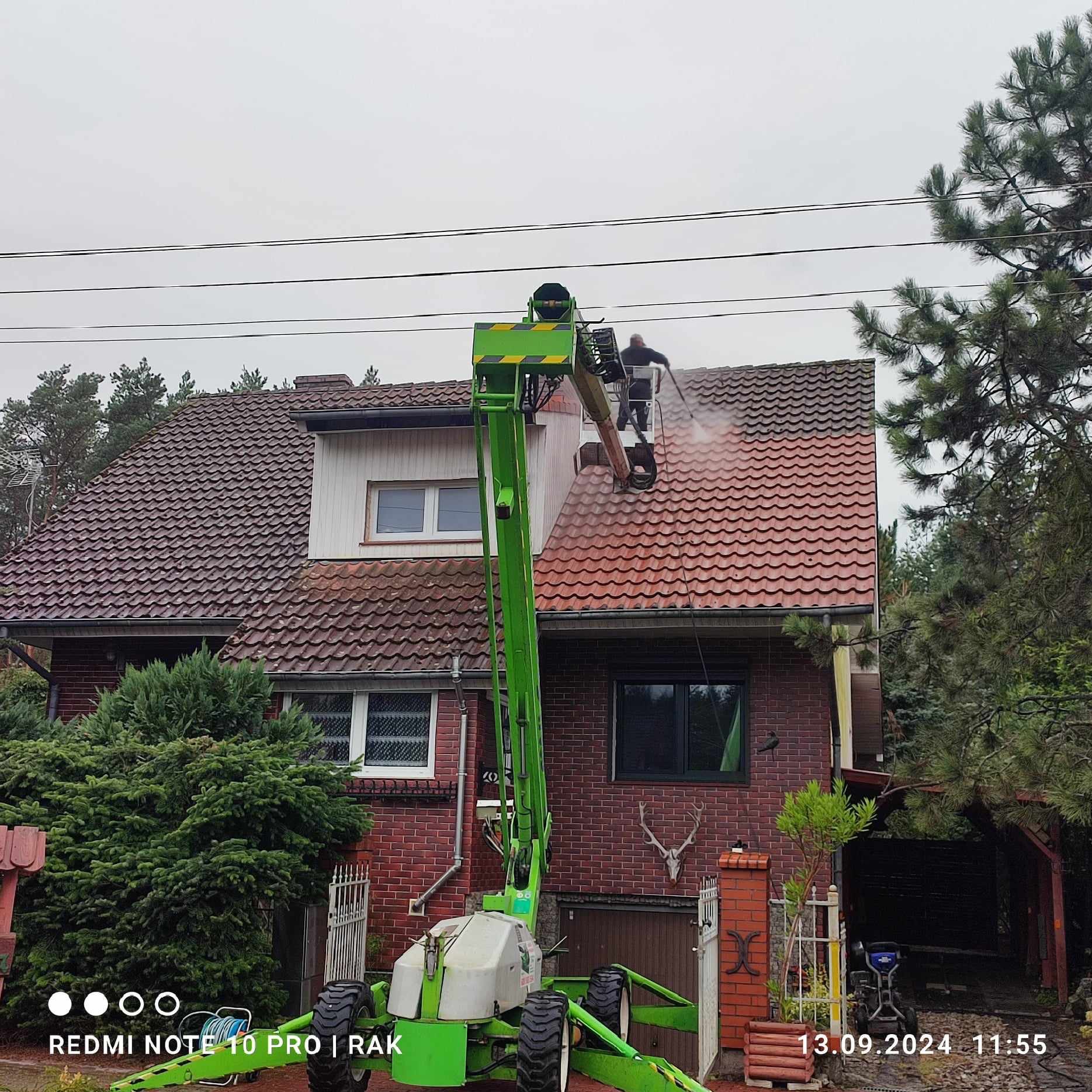 Pracownik na podnośniku koszowym czyści dach domu jednorodzinnego w Rzepinie za pomocą myjki ciśnieniowej, widoczne efekty czyszczenia.
