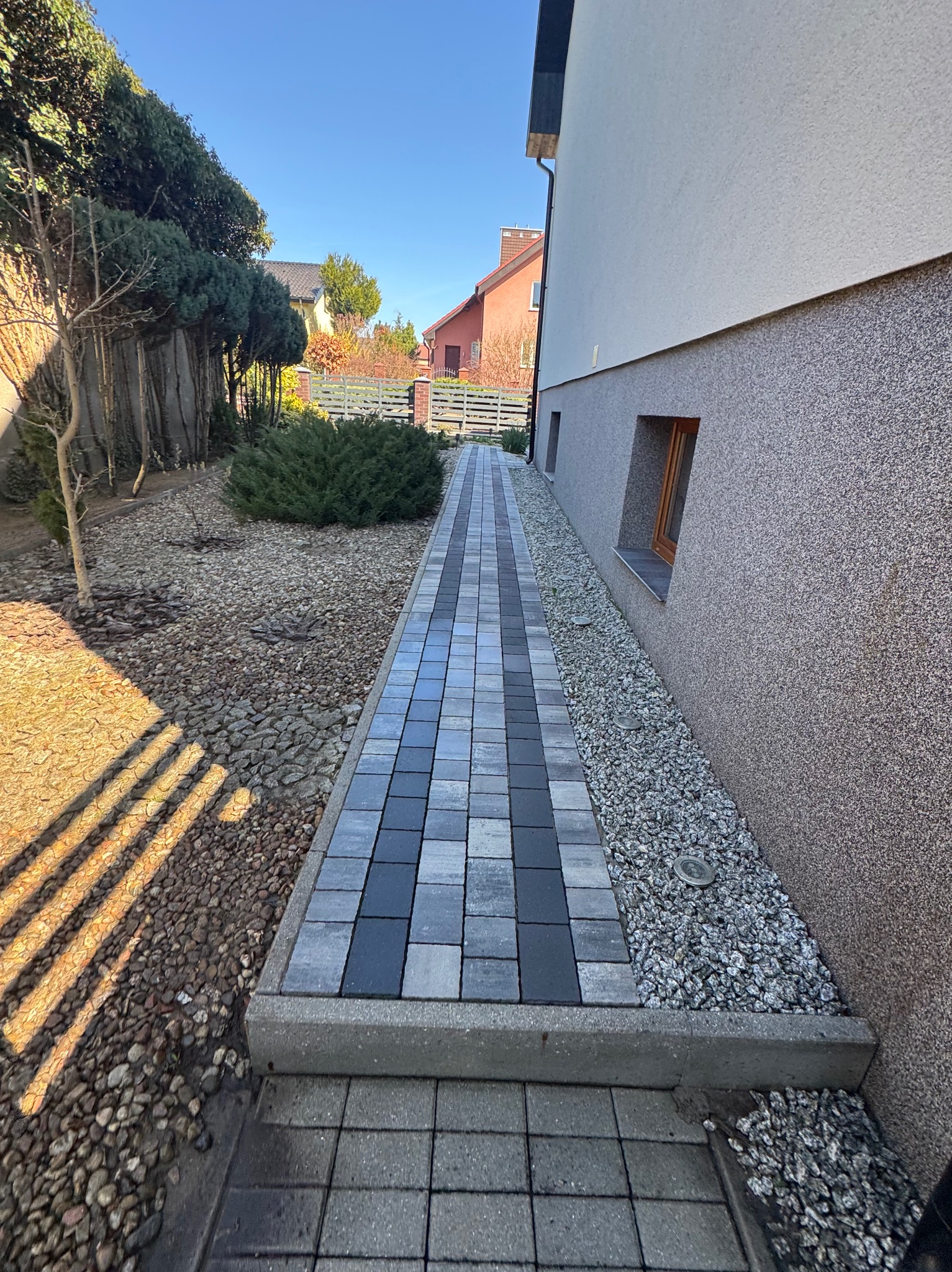 Ścieżka z kostki brukowej w odcieniach szarości prowadząca wzdłuż ściany domu z oknem, obok rabata wysypana kamieniami i krzewami, w tle inne domy.