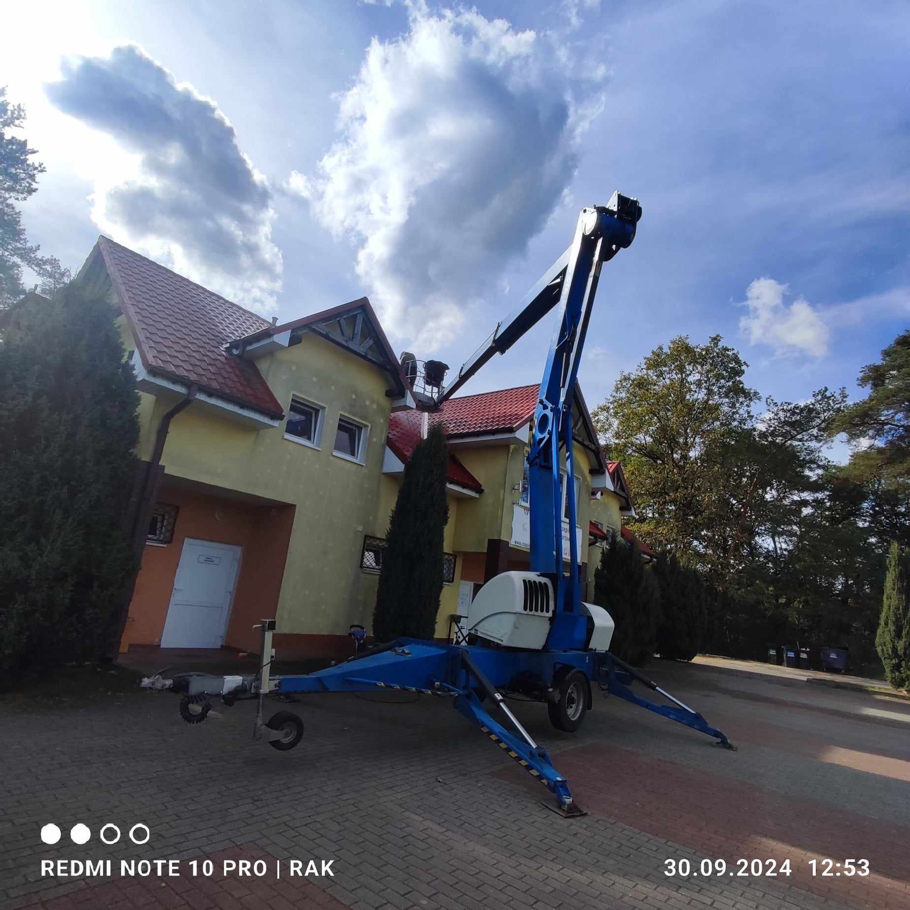 Żółty budynek z czerwoną dachówką widziany z poziomu kostki brukowej, z podnośnikiem koszowym na pierwszym planie i pracownikami na dachu.