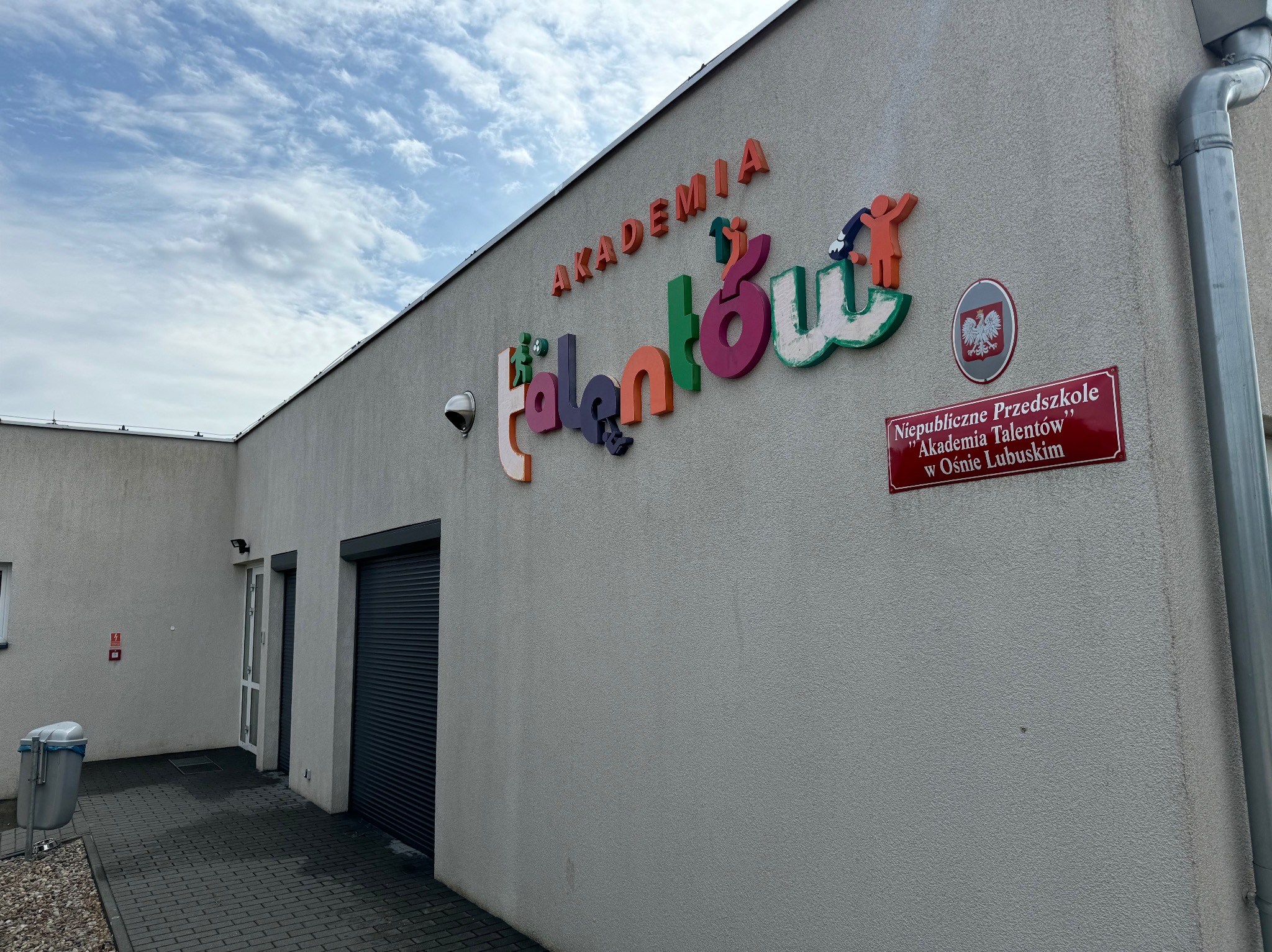 Elewacja budynku z kolorowym napisem 'Akademia Talentów' i tabliczką informacyjną 'Niepubliczne Przedszkole Akademia Talentów w Osnie Lubuskim', widoczny fragment rynny i chodnik z kostki brukowej.